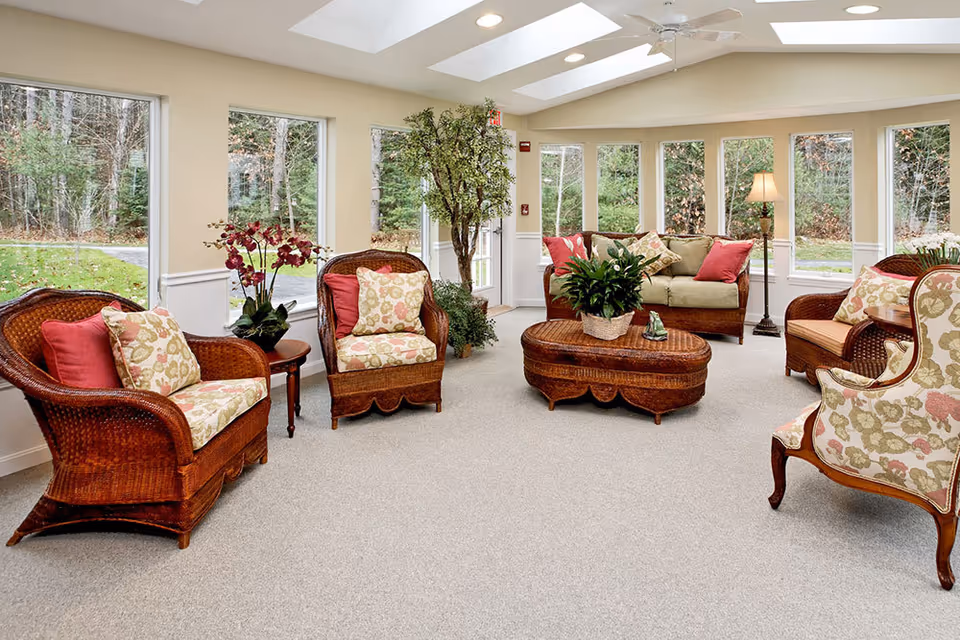 Sunlit common living room with wicker chairs and sofa, floral cushions, a central coffee table, large windows and indoor plants.