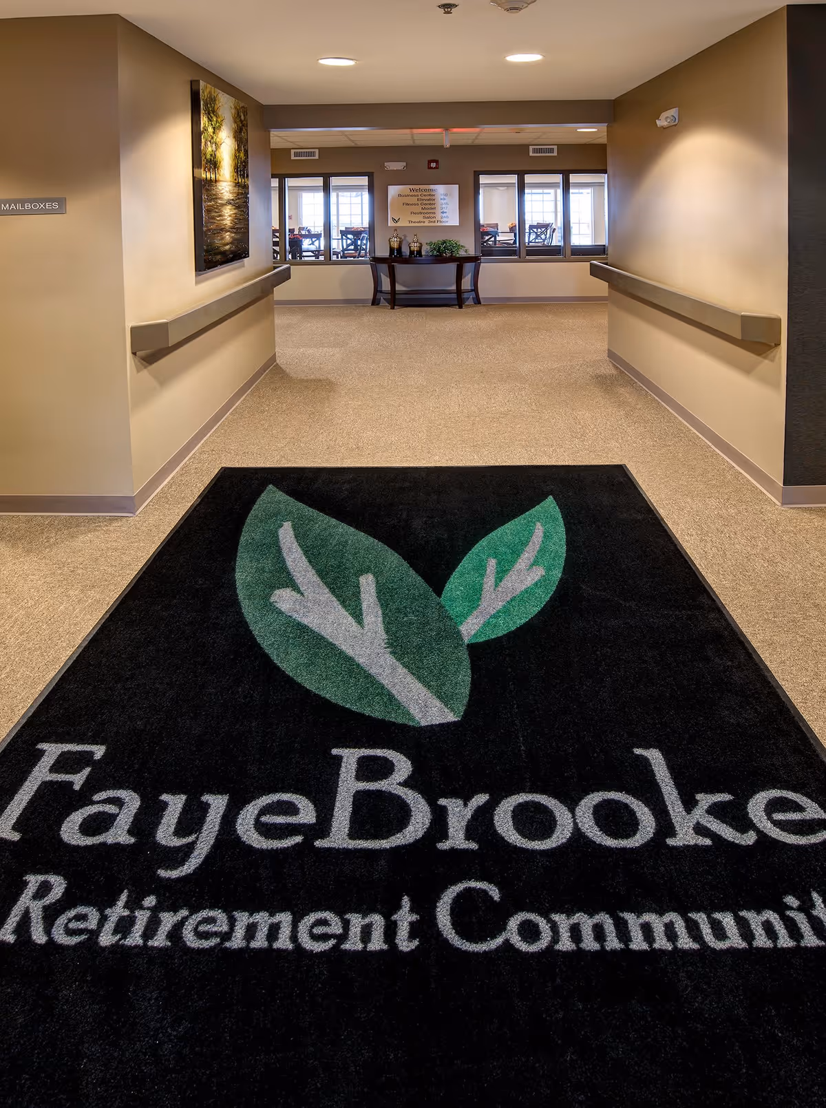 Interior hallway of Fayebrooke Retirement Community with beige walls and carpeted floor. A large black floor mat with the Fayebrooke logo featuring two green leaves is centered in the foreground. At the end of the hallway, there is a small table with decorative items and a welcome sign on the wall behind it. Windows reveal a dining area with tables and chairs beyond the hallway.