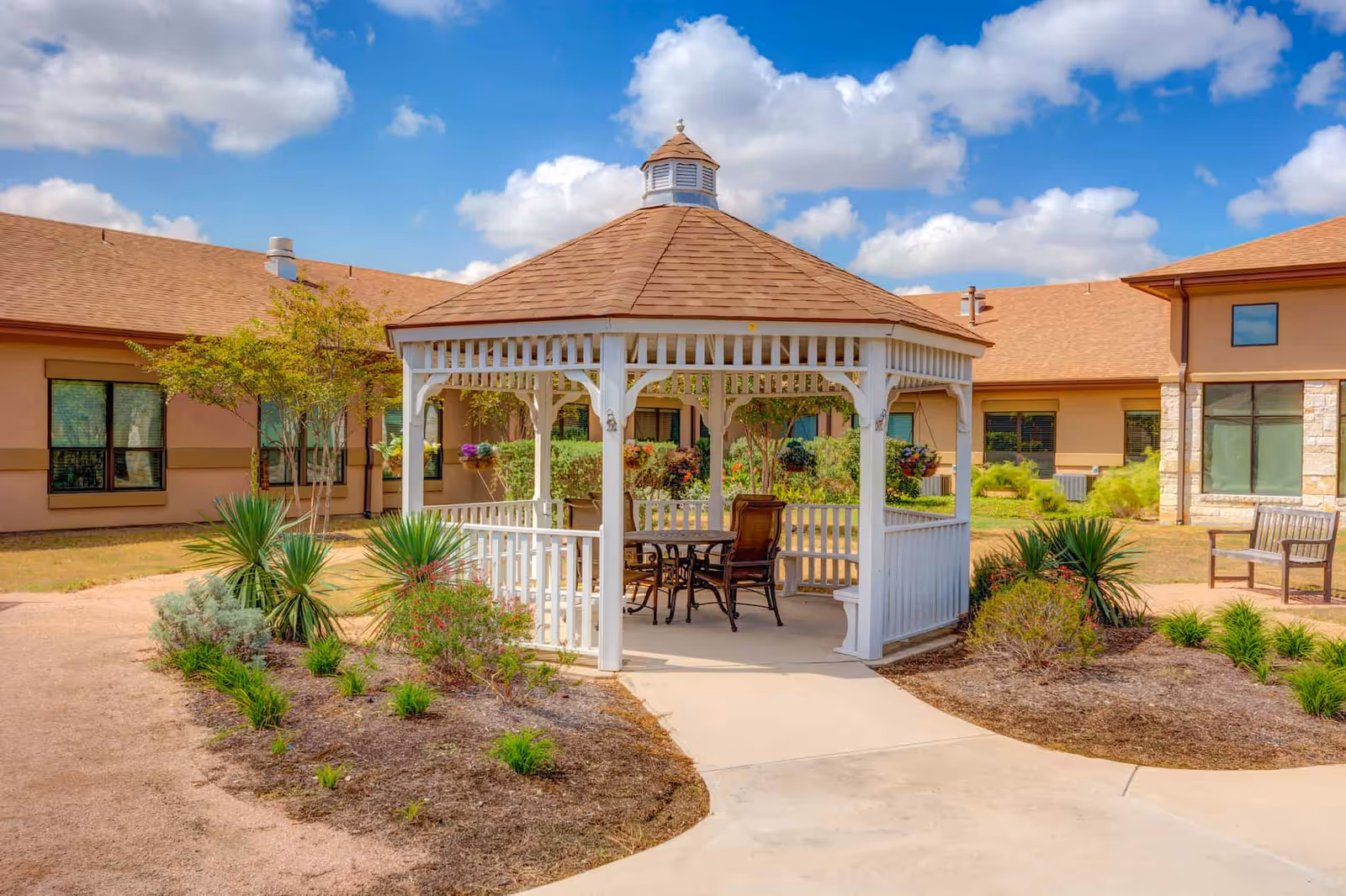 A white wooden gazebo with a brown shingled roof situated in a landscaped courtyard with plants and shrubs. The gazebo contains a table and chairs. Surrounding the courtyard are beige buildings with large windows under a blue sky with scattered clouds.