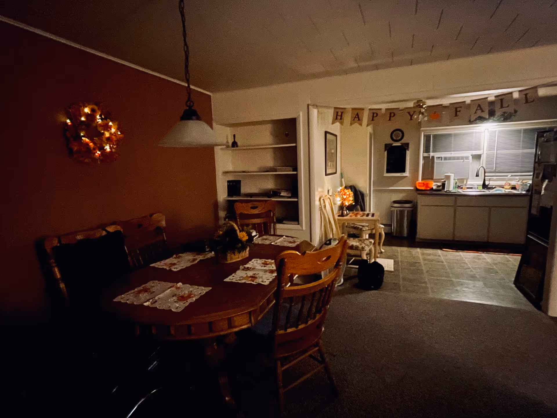 Dimly lit dining room with a wooden table, placemats, and a kitchen in the background.