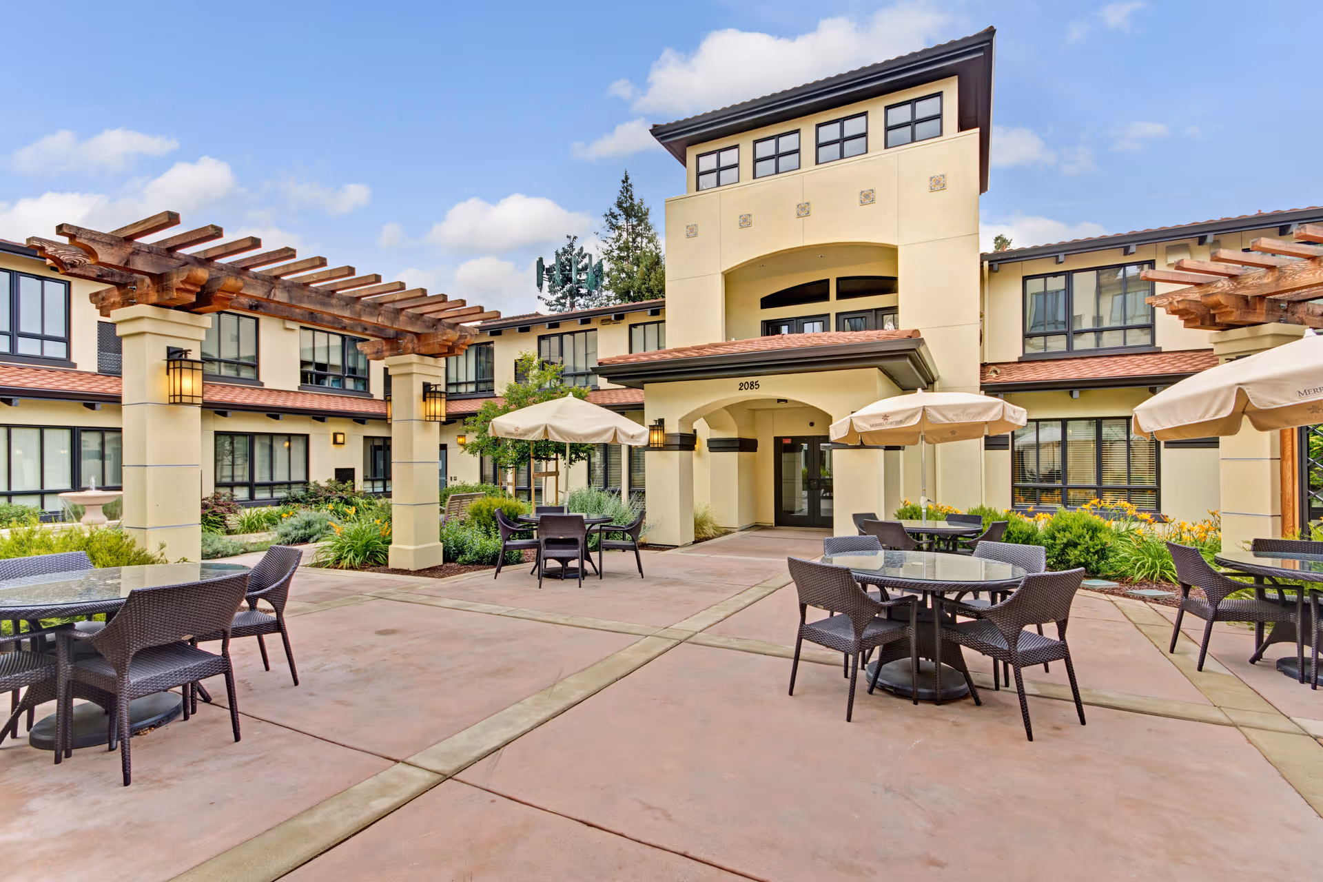 Outdoor courtyard area of Merrill Gardens at Campbell featuring patio tables with umbrellas, wicker chairs, pergolas, and landscaped greenery surrounding the seating area. The building exterior is visible with multiple windows and an entrance under an archway.