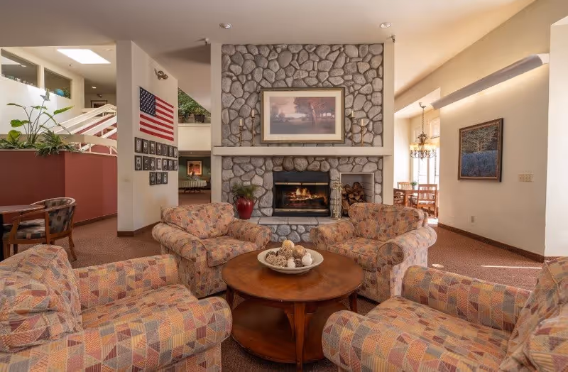 A cozy senior living common area with four patterned armchairs arranged around a round wooden coffee table with decorative items. A stone fireplace with a framed painting above it is centered in the background. To the left, there is an American flag and framed photos on the wall. A dining area with a chandelier is visible in the background to the right.