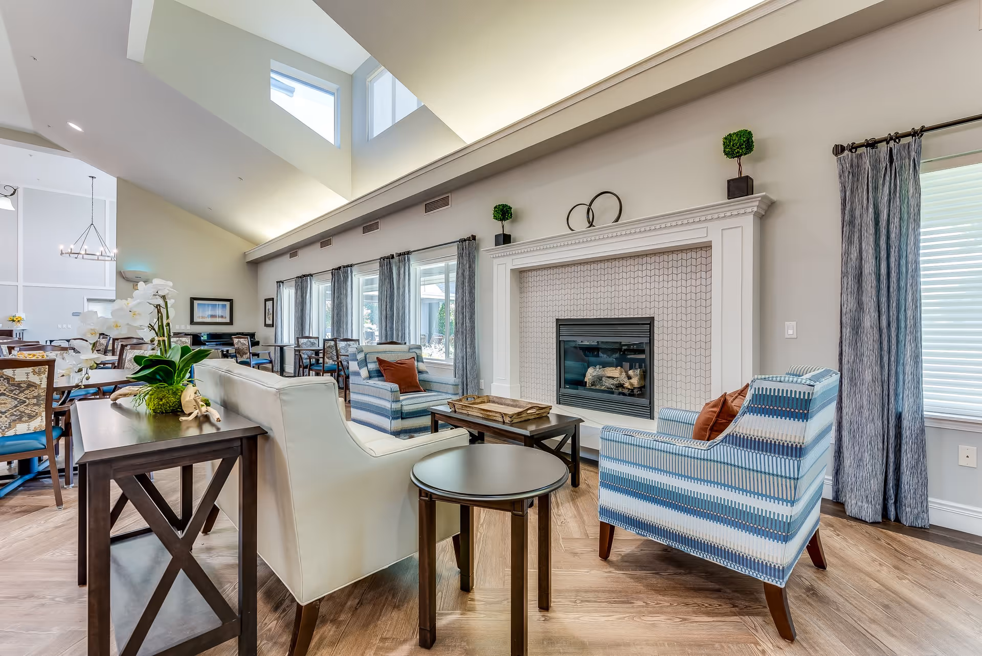 A bright and spacious senior living common area with high vaulted ceilings and large windows. The room features a white fireplace with decorative tiles, two blue patterned armchairs with orange cushions, a white sofa, and dark wood tables. In the background, there is a dining area with tables and chairs, and the space is decorated with plants and curtains.