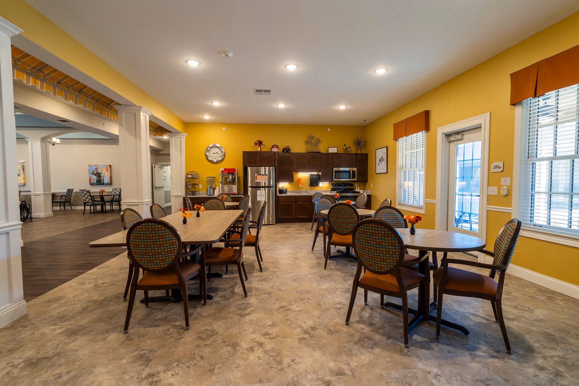 A bright dining area with yellow walls and multiple tables and chairs arranged for seating. The room features a kitchenette with a refrigerator, microwave, and cabinets along the back wall. There are windows with blinds and orange valances letting in natural light. Small flower arrangements are placed on the tables.