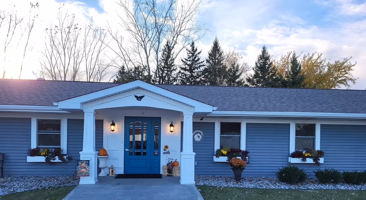 Single-story blue building front with a covered white-column entrance, teal double doors, and fall decorations including pumpkins and potted flowers.