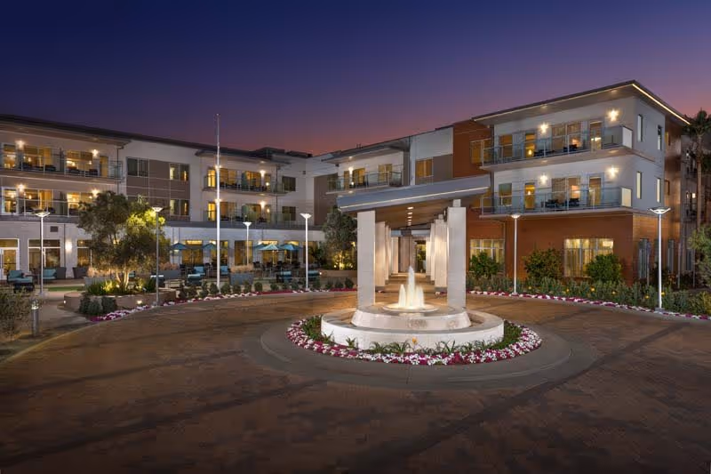Exterior view of a modern senior living facility at dusk with a circular driveway featuring a central water fountain surrounded by flowers. The building has multiple floors with balconies and large windows, and outdoor seating areas are visible. The sky is a gradient of purple and orange as evening sets in.