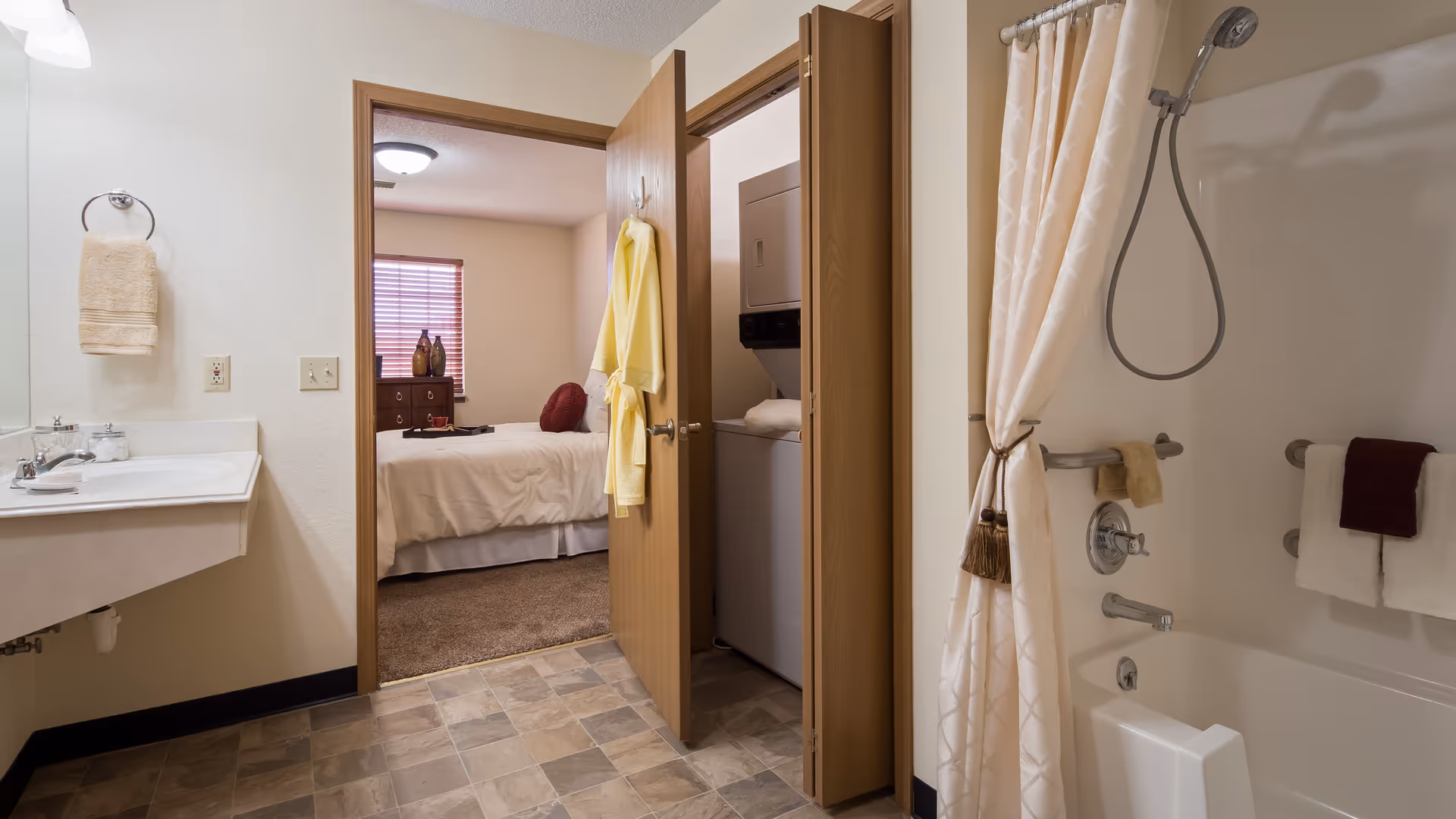Bathroom with a white sink and beige towel on a ring, a bathtub with a showerhead and a white shower curtain tied back, and a door leading to a bedroom with a bed and a window with blinds. Another door reveals a laundry area with a stacked washer and dryer. A yellow bathrobe hangs on the bathroom door.