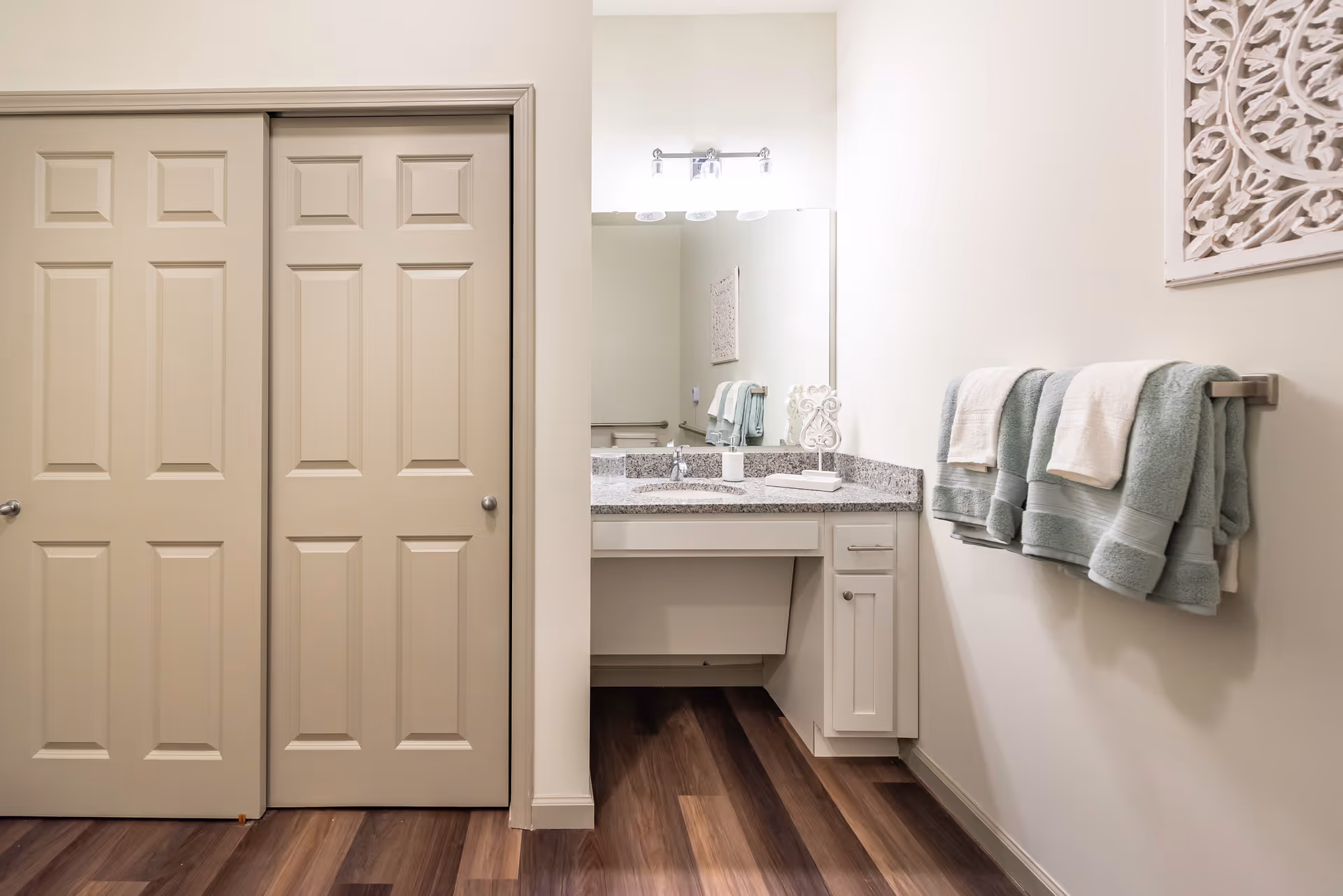 A bathroom vanity area with a granite countertop, a large mirror, and a light fixture above. There are neatly folded towels hanging on a towel rack on the right wall, and a decorative white wall art piece above the towels. To the left, there are two beige sliding closet doors. The floor is wood with a dark finish.