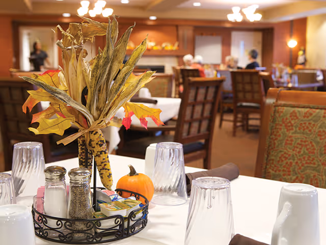 A dining table with a fall-themed centerpiece, condiments, and glasses in a restaurant-style room with chairs and people in the background.