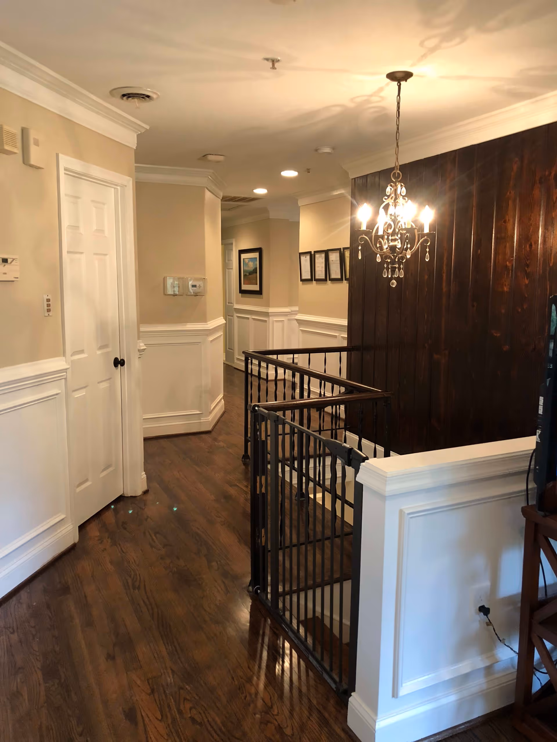Interior hallway with dark hardwood floors, white wainscoting, a chandelier, and a gated staircase.