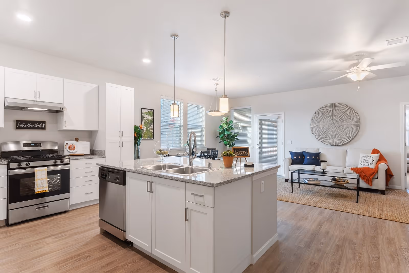 A bright and modern open-concept kitchen and living room area. The kitchen features white cabinets, a stainless steel stove and dishwasher, a large island with a double sink, and pendant lights hanging above. The living room has a white sofa with blue and orange pillows, a glass coffee table, a large circular wall decoration, and a ceiling fan. There are windows with blinds and a door leading outside.