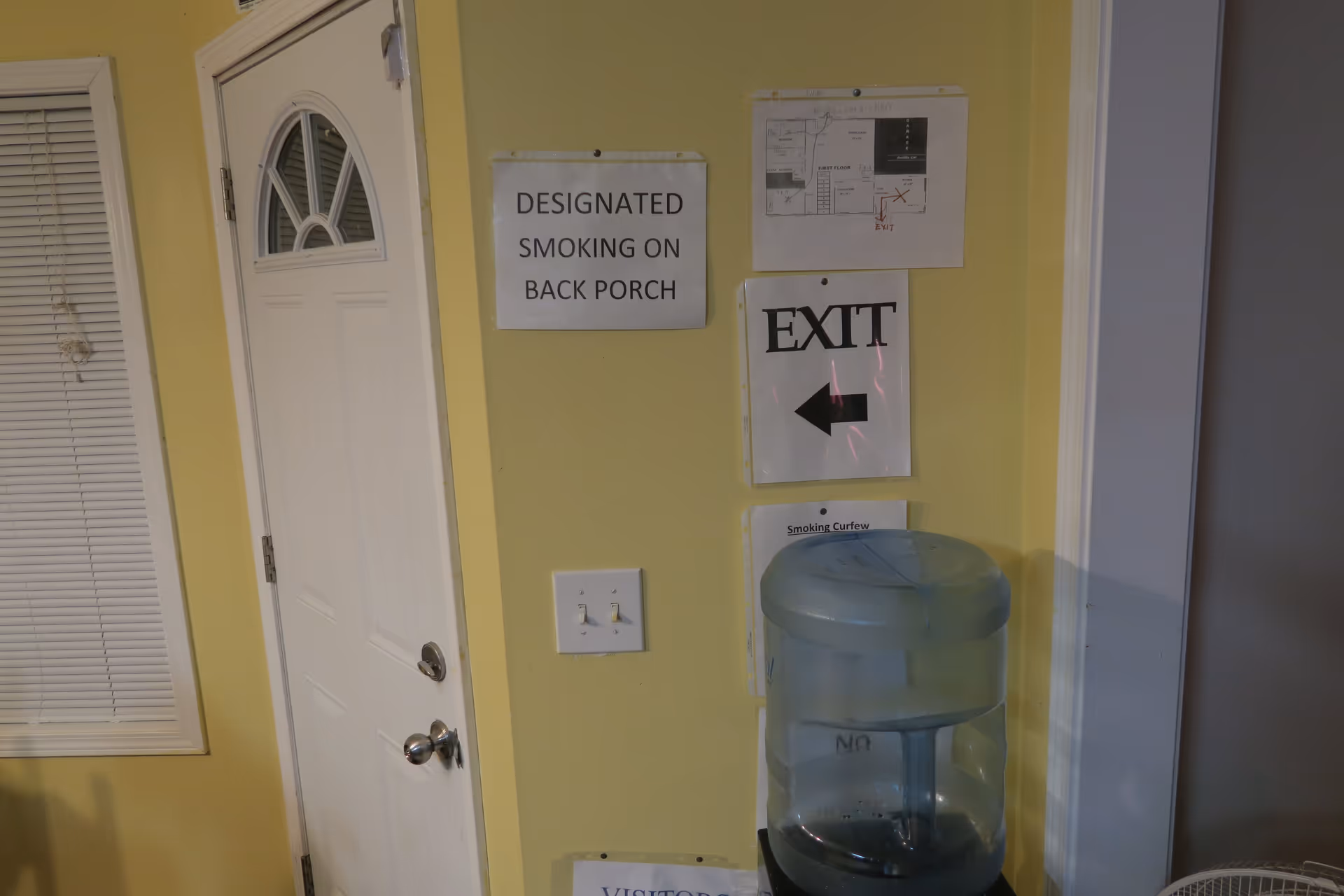 Interior corner of a room with a white door and yellow walls. Several signs are posted on the wall, including one that says 'DESIGNATED SMOKING ON BACK PORCH,' another with a floor plan, an 'EXIT' sign with a left arrow, and a partially visible 'Smoking Curfew' notice. A water cooler is positioned against the wall below the signs.