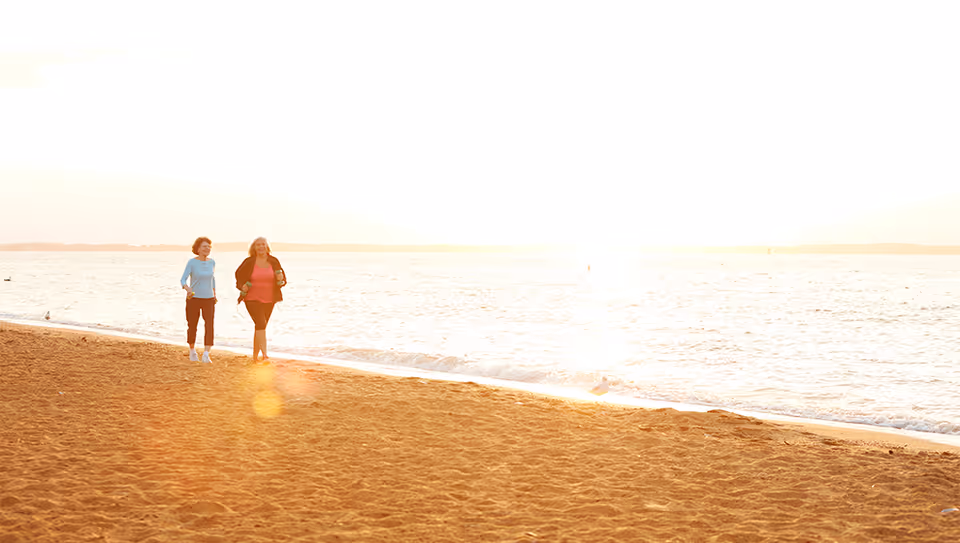 Two women walking and enjoying a sunny day on a sandy beach near the water's edge with gentle waves and a bright sky.