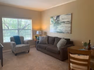 A cozy living room with a large window letting in natural light. The room features a gray sofa with two cushions, a patterned armchair, a small wooden dining table with two chairs, a table lamp on a side table, and a large abstract painting on the beige wall.