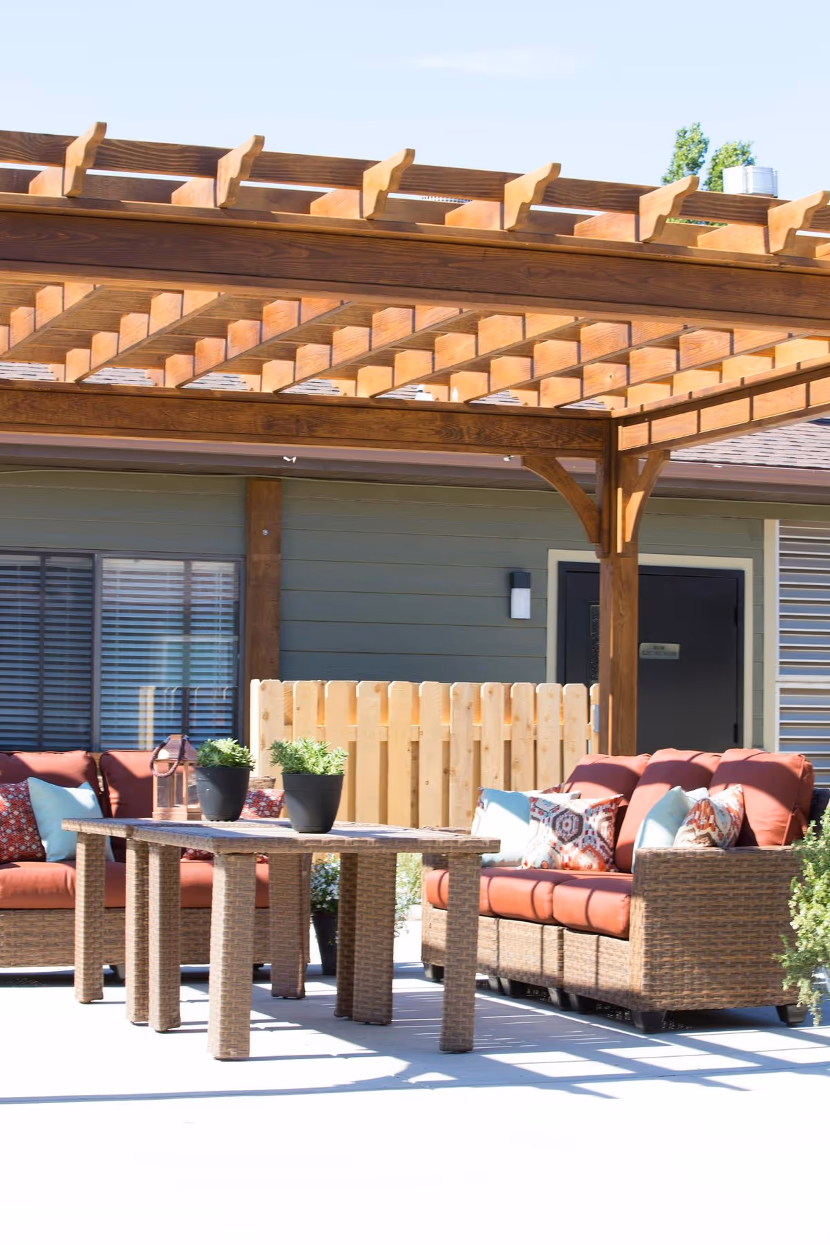 Outdoor seating area with two wicker sofas with red cushions and decorative pillows under a wooden pergola. A rectangular wicker table with potted plants is placed between the sofas. The background shows a building with green siding, a window with blinds, a wooden fence, and a door.