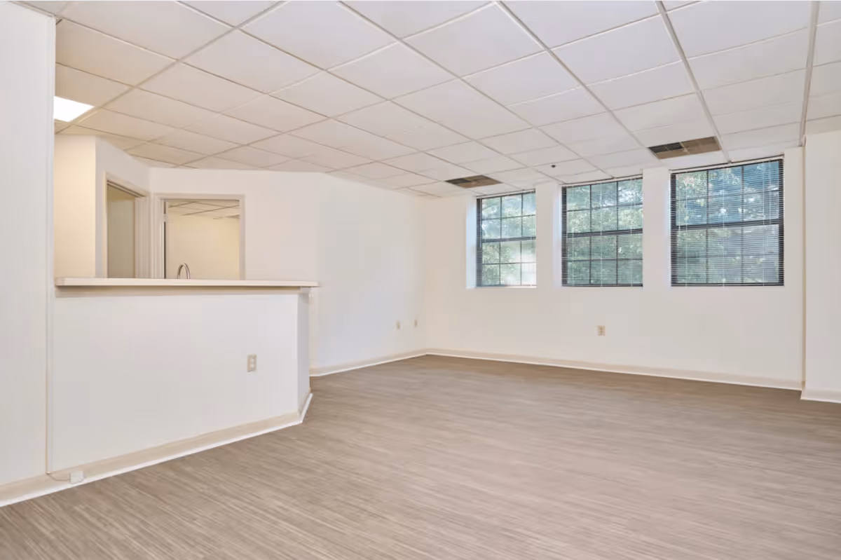 Empty room with light-colored walls and wood-patterned flooring, featuring three large windows with blinds and a partial view of a kitchen area with a counter and sink.