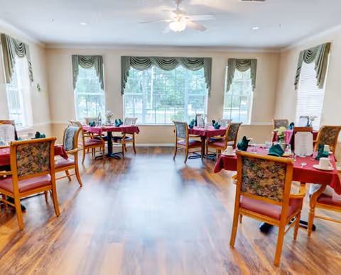 A bright dining room with wooden floors and several tables covered with red tablecloths. Each table is set with green napkins, white cups, and silverware. The room has large windows with green valances, allowing natural light to fill the space. Wooden chairs with floral upholstery surround the tables.