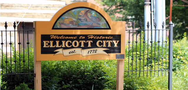 Historic Ellicott City welcome sign with arch, motto, and iron fence.