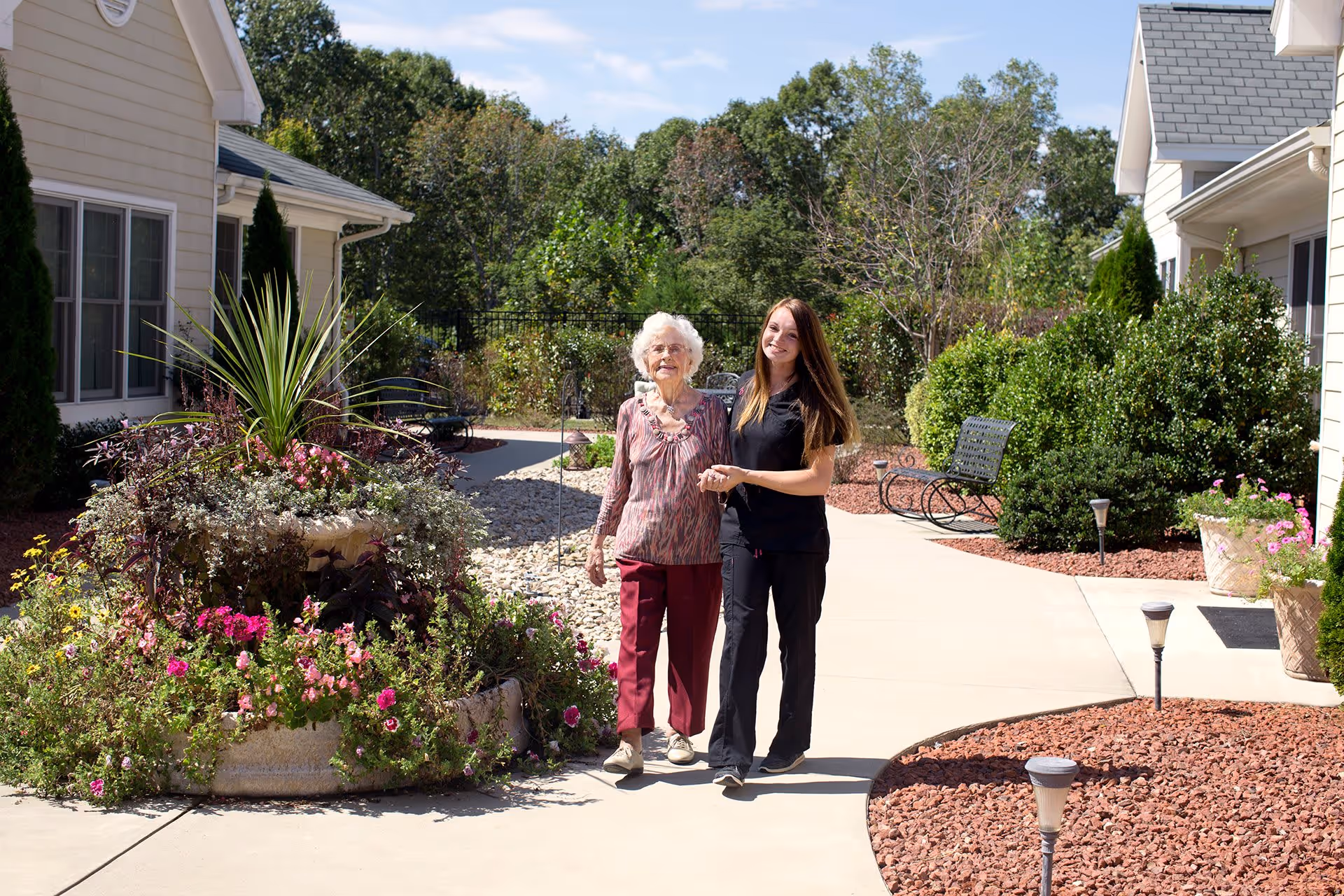 An elderly woman and a younger woman walking together on a paved pathway in a garden area between two buildings. The garden has various plants, flowers, and shrubs, with benches and outdoor lighting along the path. The sky is clear and sunny.