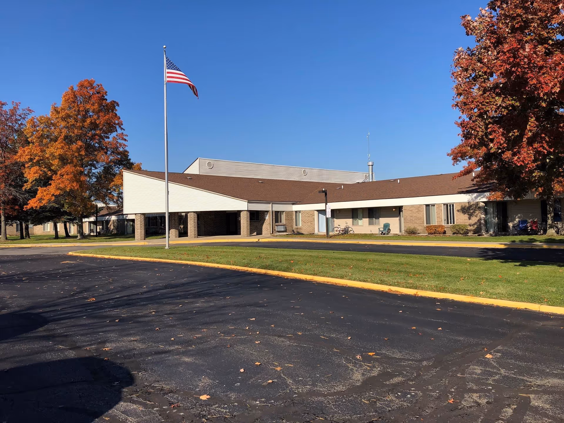 Exterior view of a single-story building with a brown roof and beige siding, surrounded by green grass and trees with autumn-colored leaves. An American flag is flying on a flagpole in front of the building under a clear blue sky.