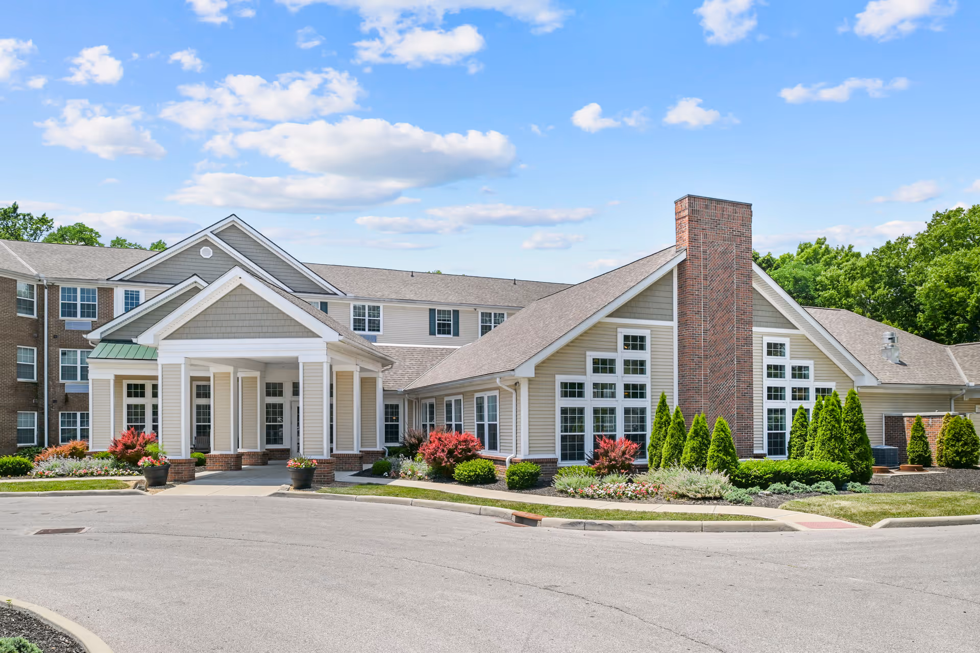 Front exterior of a senior living building with a covered portico entrance, large windows, brick chimney, and landscaped grounds under a blue sky.