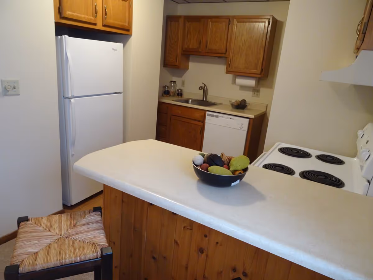 A small kitchen with wooden cabinets, a white refrigerator, a white electric stove with four coil burners, a dishwasher, and a sink. There is a countertop with a bowl of assorted decorative fruits and a wooden stool with a woven seat in front of it.
