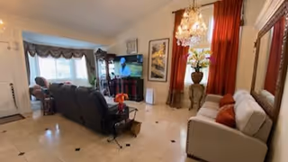 A spacious living room with beige tiled floor, a white sofa with red cushions on the right, a dark reclining sofa facing a TV mounted on a wooden cabinet, large windows with curtains, a chandelier hanging from the ceiling, and decorative plants and framed pictures on the walls.