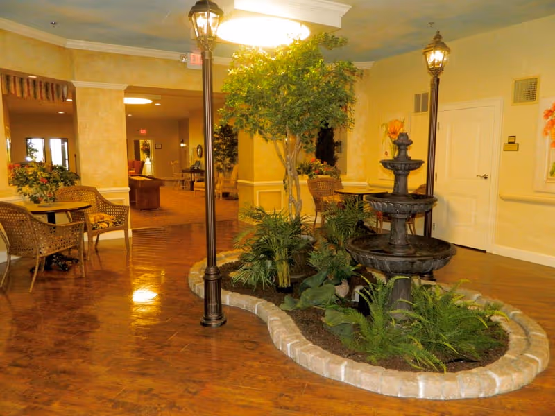 Indoor communal seating area with a central tiered fountain, plants, wicker chairs and lamp posts in a warmly decorated lobby.