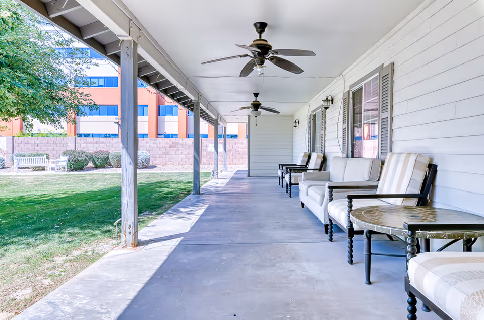 Covered outdoor patio area with ceiling fans, cushioned chairs, and a round table. The patio overlooks a grassy area with bushes, trees, and benches. A building with windows and a brick wall is visible in the background.