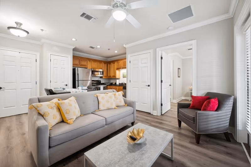 A bright and modern living room area with a gray sofa adorned with yellow and white patterned pillows, a gray armchair with red pillows, and a light-colored coffee table with a decorative bowl. The room has wood flooring, white walls, and a ceiling fan with a light. In the background, there is a kitchen with wooden cabinets, stainless steel appliances, and a dining table with chairs. Several white doors lead to other rooms.