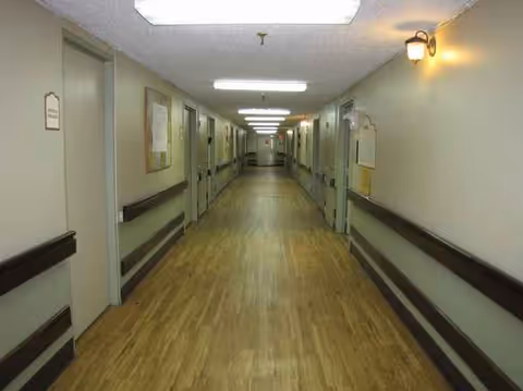 Long, empty hallway in a nursing facility with doors, handrails, and overhead lights.