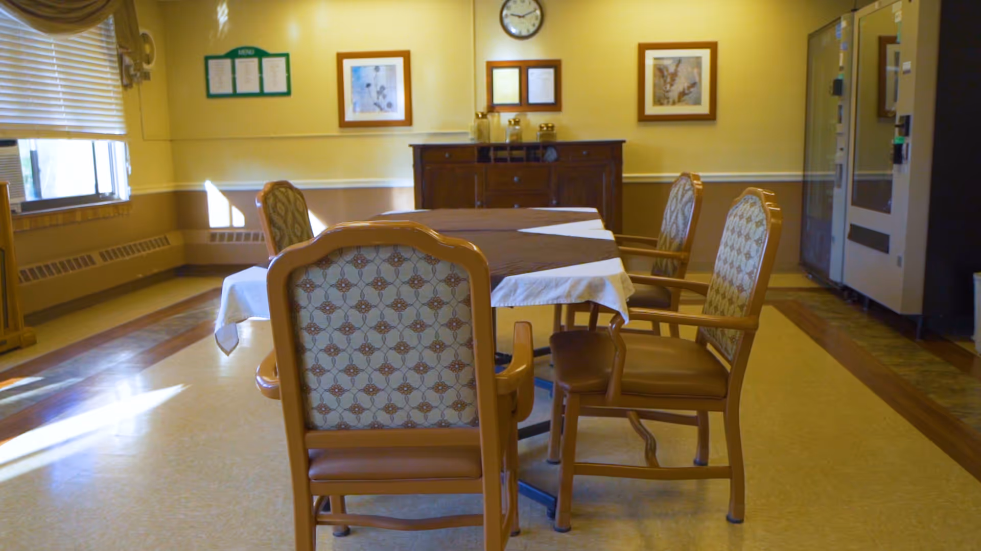 Dining room with a table covered by a tablecloth surrounded by padded wooden chairs in a bright room