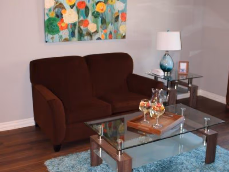 A cozy living room area with a dark brown loveseat, a glass coffee table with a wooden tray holding three glasses of a beverage, a blue area rug, a side table with a blue glass lamp, a small framed photo, and a colorful floral painting on the wall.