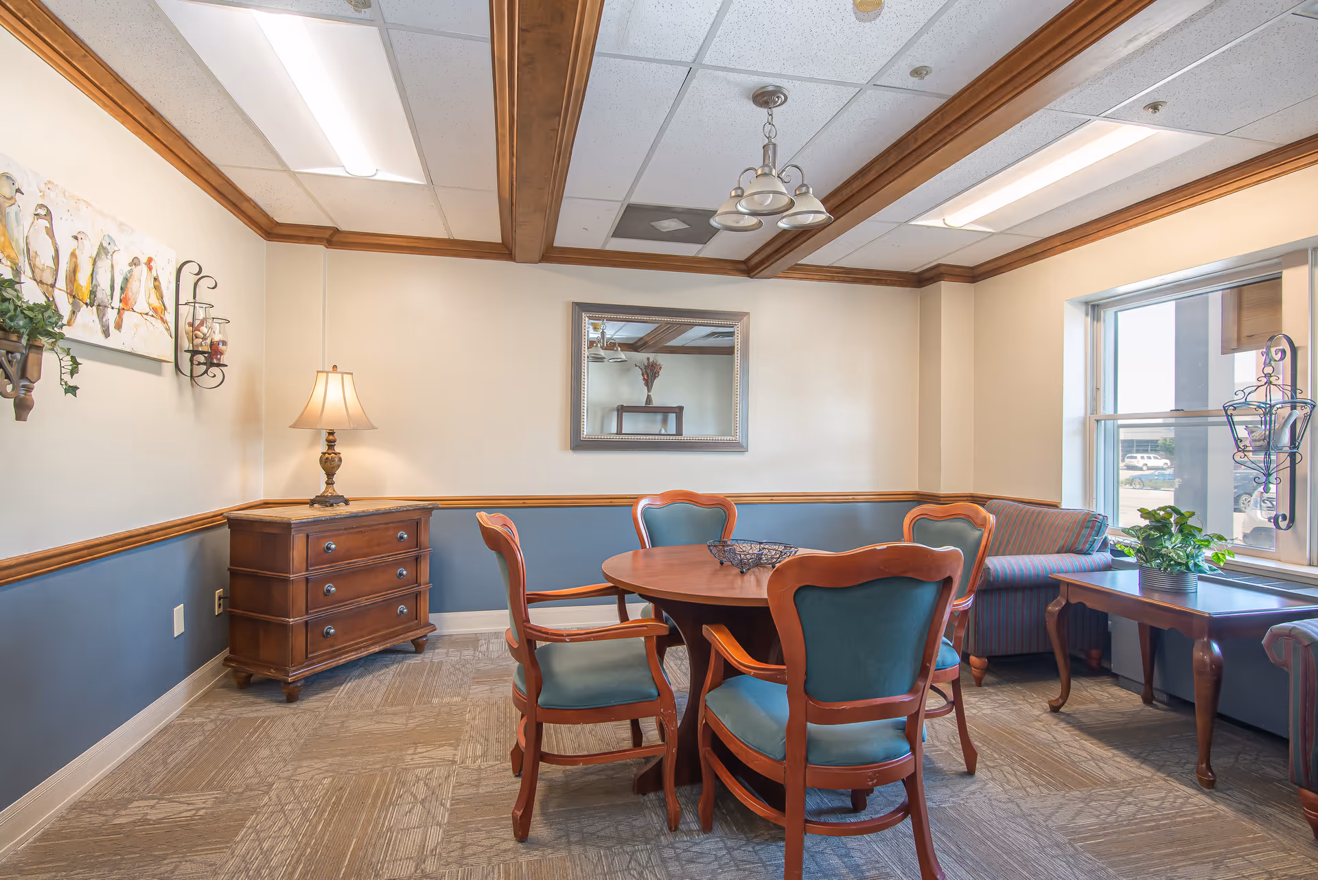 A cozy interior room with a round wooden table surrounded by four wooden chairs with teal cushions. There is a wooden chest of drawers with a lamp on it against the wall, a striped sofa near a window, and a wooden side table with a plant. The walls are painted cream and blue with wooden trim, and a mirror and bird artwork hang on the walls. The ceiling has wooden beams and a light fixture.