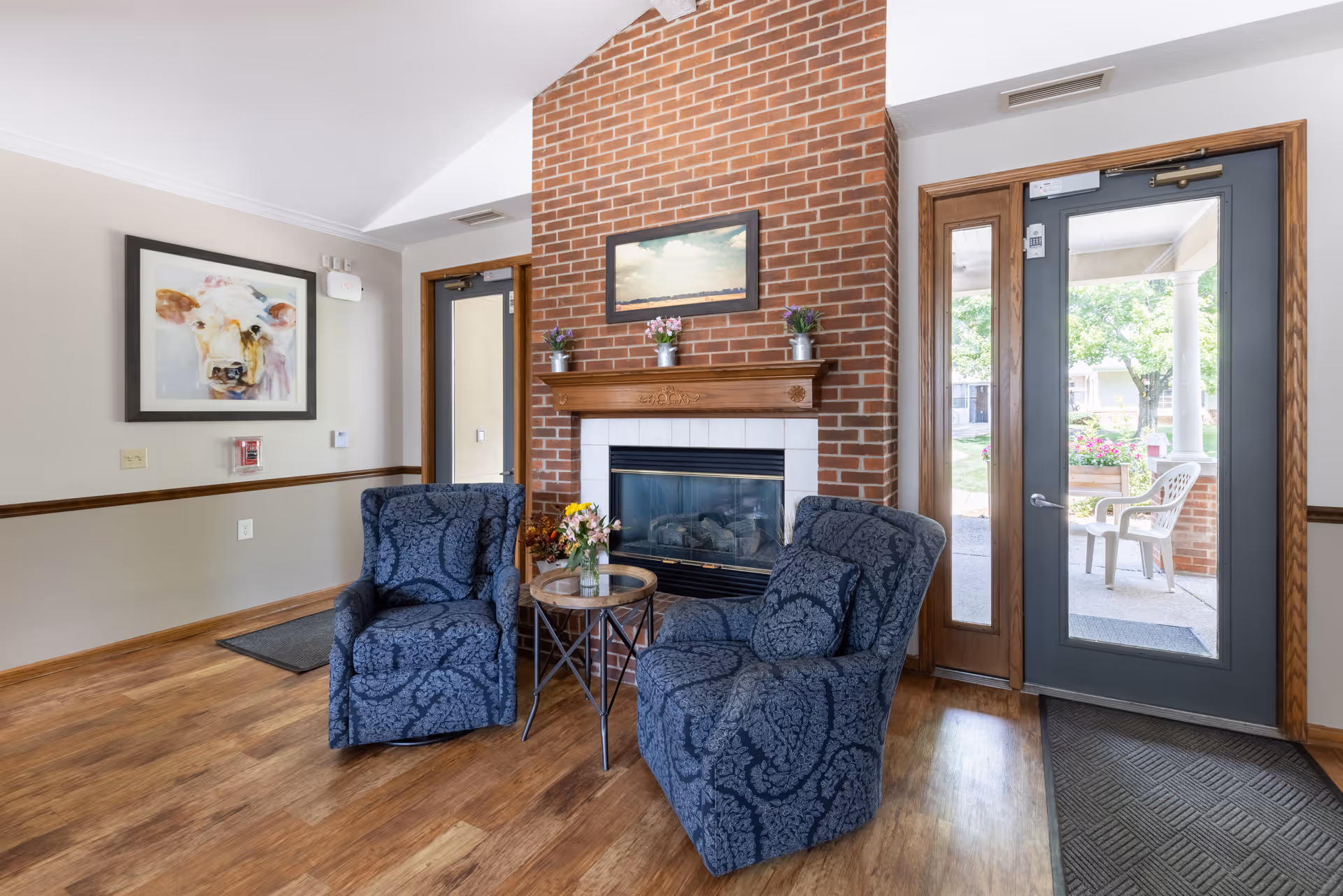 A cozy sitting area with two patterned blue armchairs, a small round table, and a brick fireplace next to a glass exterior door.