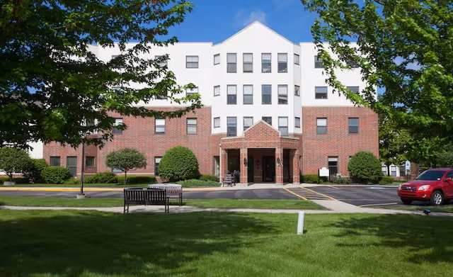 Front exterior view of a multi-story senior living facility building with a brick and white facade, surrounded by green trees and a well-maintained lawn. There is a bench on the grass and a red vehicle parked on the right side near the entrance.