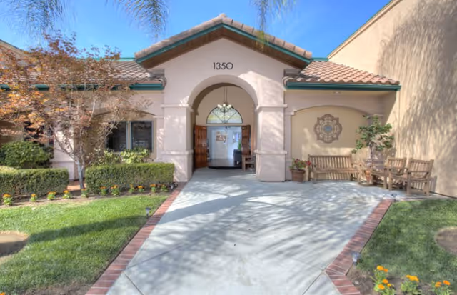 Front exterior view of a single-story building with a tiled roof and an arched entrance. The building number 1350 is displayed above the entrance. There are green bushes, a tree, and a small garden area with flowers on either side of the concrete walkway leading to the entrance. On the right side, there are wooden benches and potted plants against the building wall.