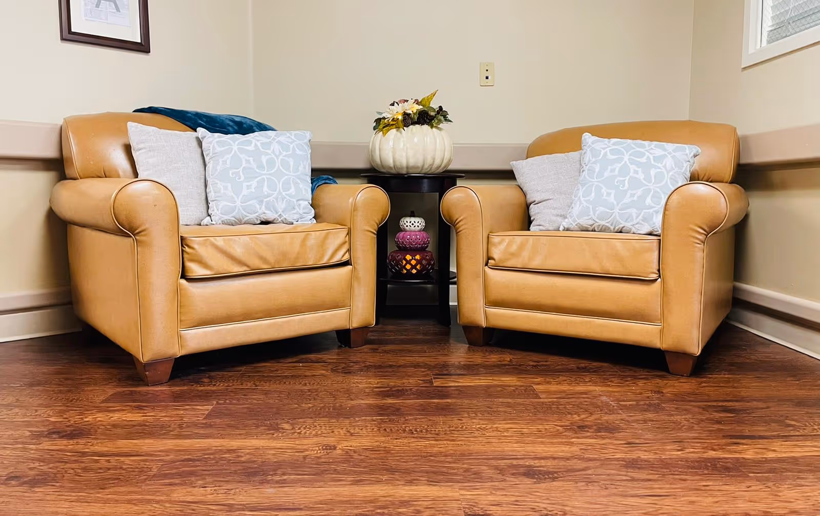 Two tan leather armchairs with decorative pillows are positioned on a wooden floor with a small black side table between them holding a white pumpkin-shaped vase with flowers and two decorative candle holders underneath. The room has beige walls and a window on the right side.