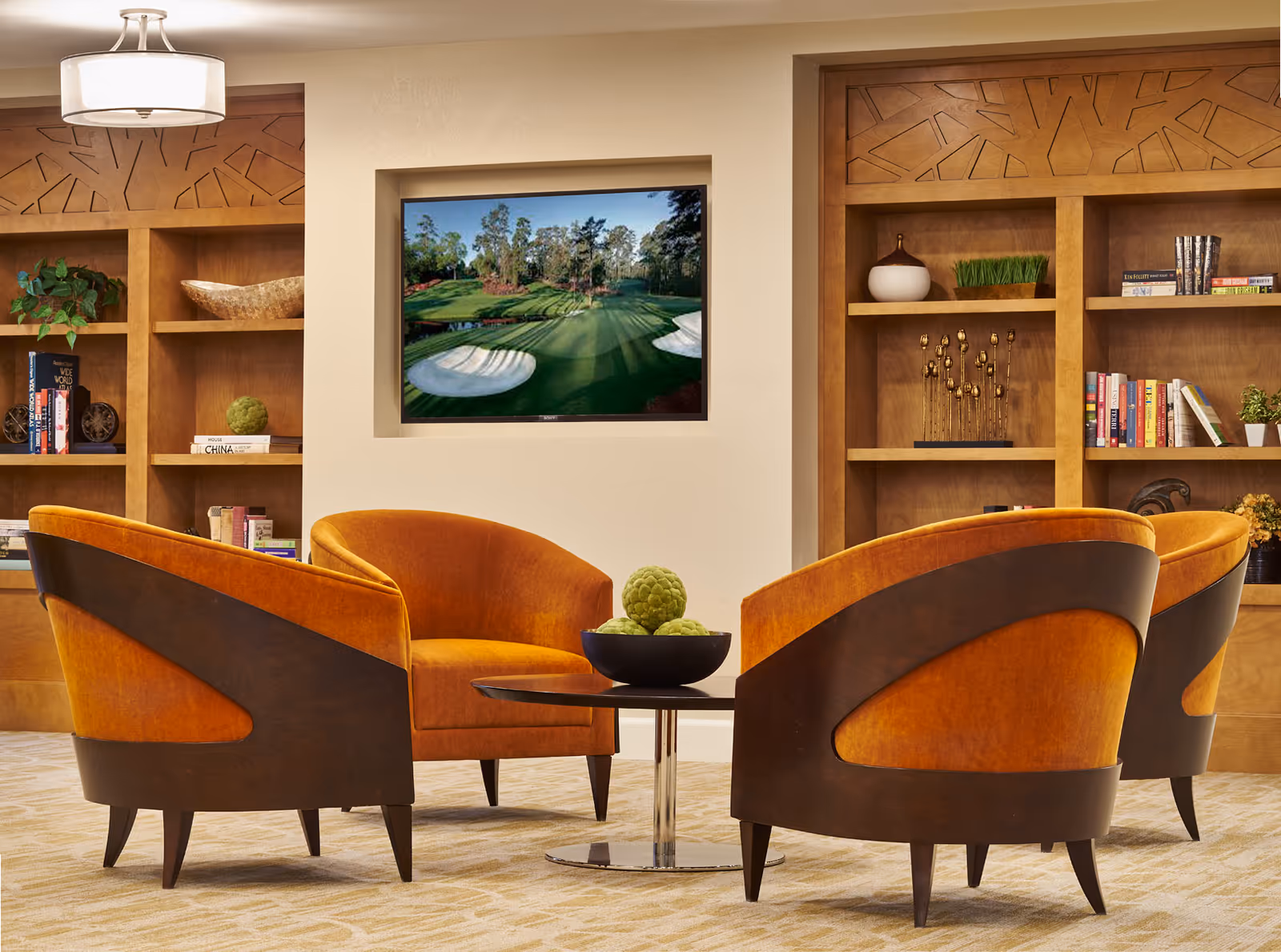 A cozy seating area with four orange upholstered chairs arranged around a small round table with a decorative bowl of green spheres. Behind the seating area are built-in wooden shelves with books, plants, and decorative items. A flat-screen TV is mounted on the wall displaying a golf course scene.