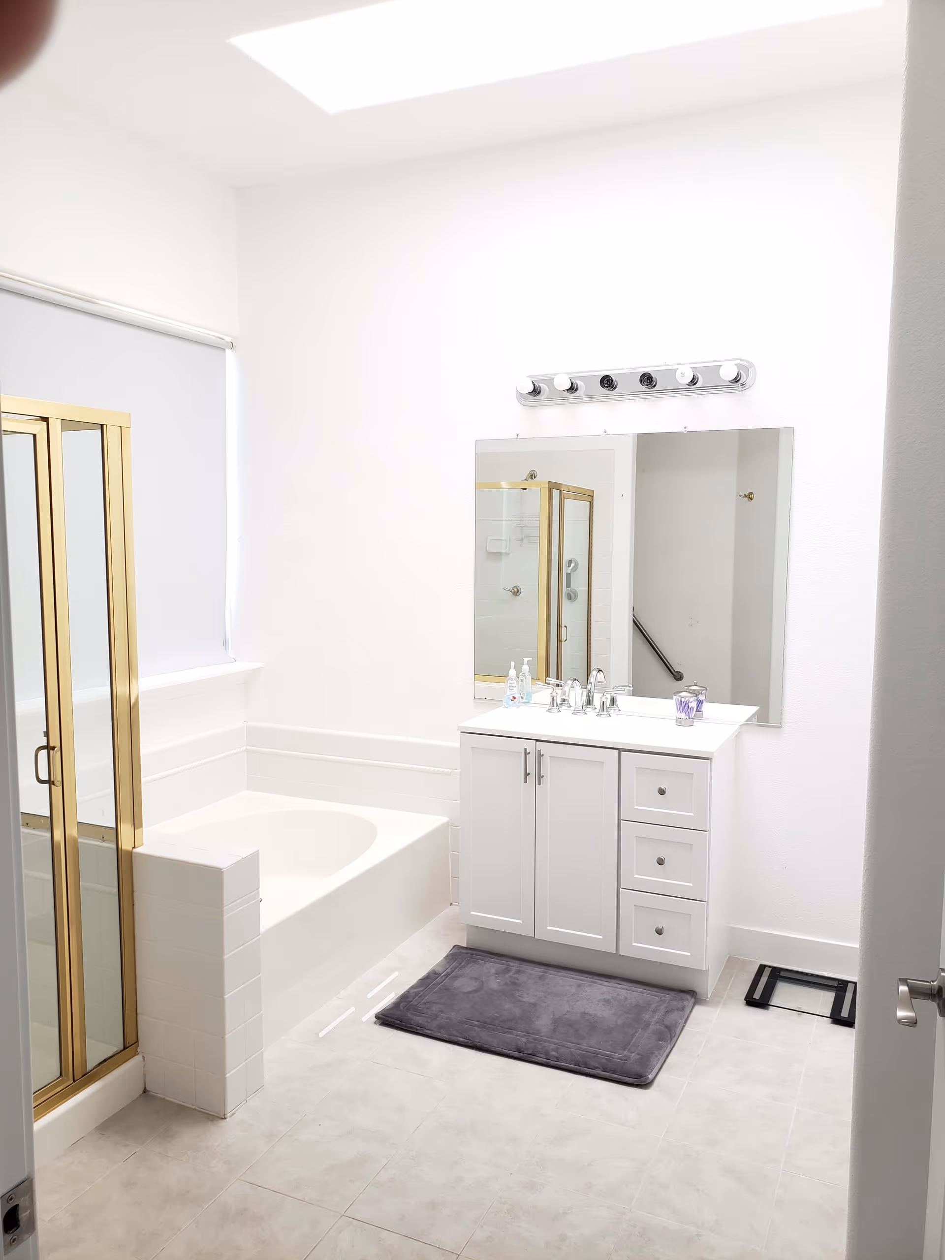 A bright and clean bathroom featuring a white bathtub, a separate shower with gold-framed glass doors, a white vanity with a sink and three drawers, a large mirror above the vanity, a gray bath mat on the tiled floor, and a digital scale near the door. The walls are white, and there is a skylight providing natural light.
