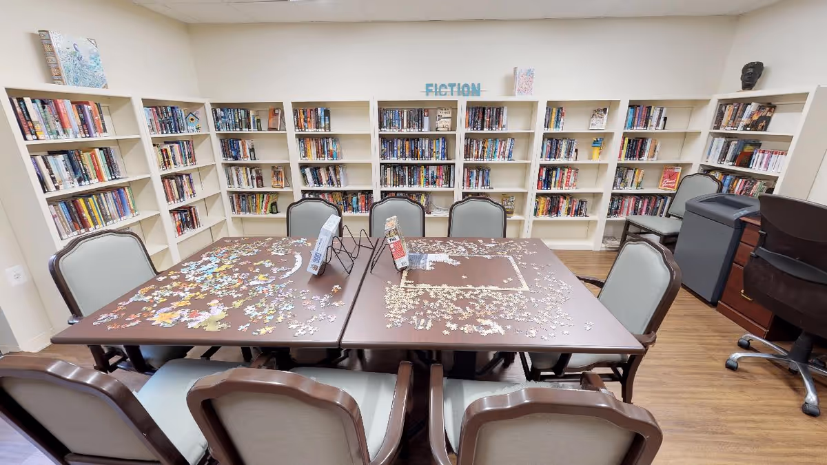 A cozy library room with white bookshelves filled with books along the walls. In the center, two tables pushed together hold partially completed jigsaw puzzles. Several cushioned chairs surround the tables. The room has wooden flooring and a sign labeled 'FICTION' on the back wall above the bookshelves.