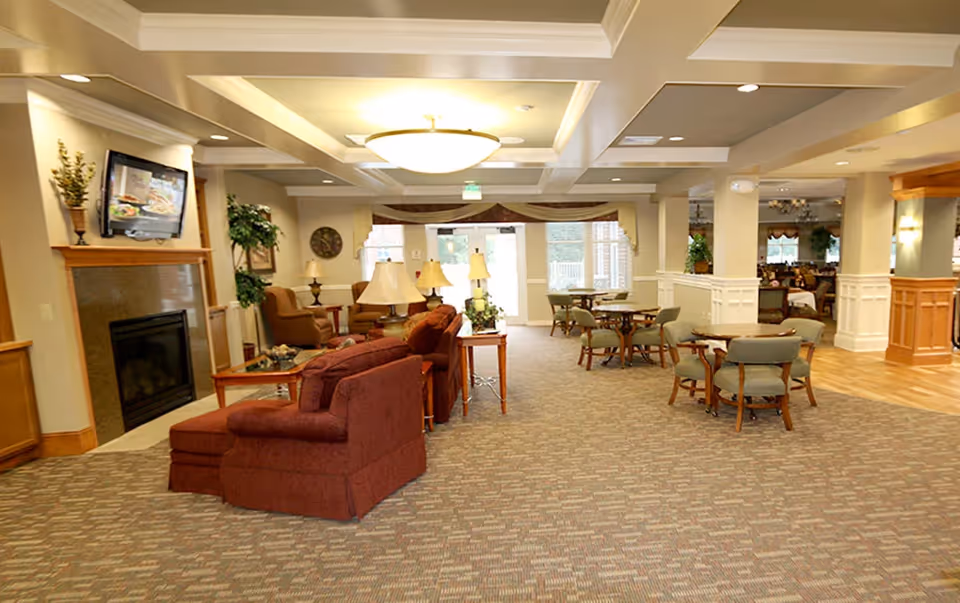 A spacious senior living common area with carpeted floor, multiple seating arrangements including red upholstered armchairs and green cushioned chairs around wooden tables. There is a fireplace with a mounted TV above it on the left side, several table lamps, potted plants, and large windows letting in natural light at the far end.