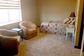 A cozy bedroom with a single bed covered in a patterned bedspread, two cushioned armchairs, a small side table, a white nightstand with a lamp, and a window with blinds letting in natural light.