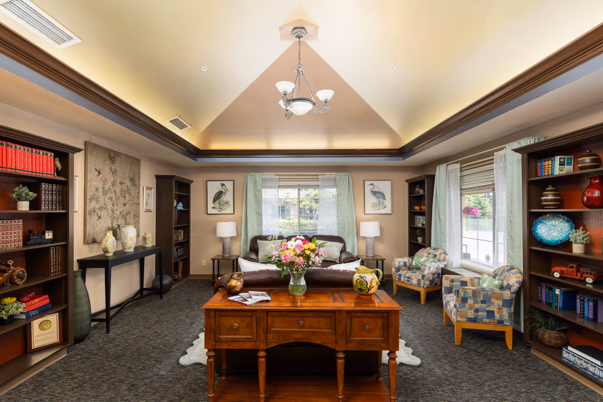 A comfortable lounge with a sofa, patterned armchairs, bookshelves, a central wooden table with a vase of flowers, and large windows under a tray ceiling.