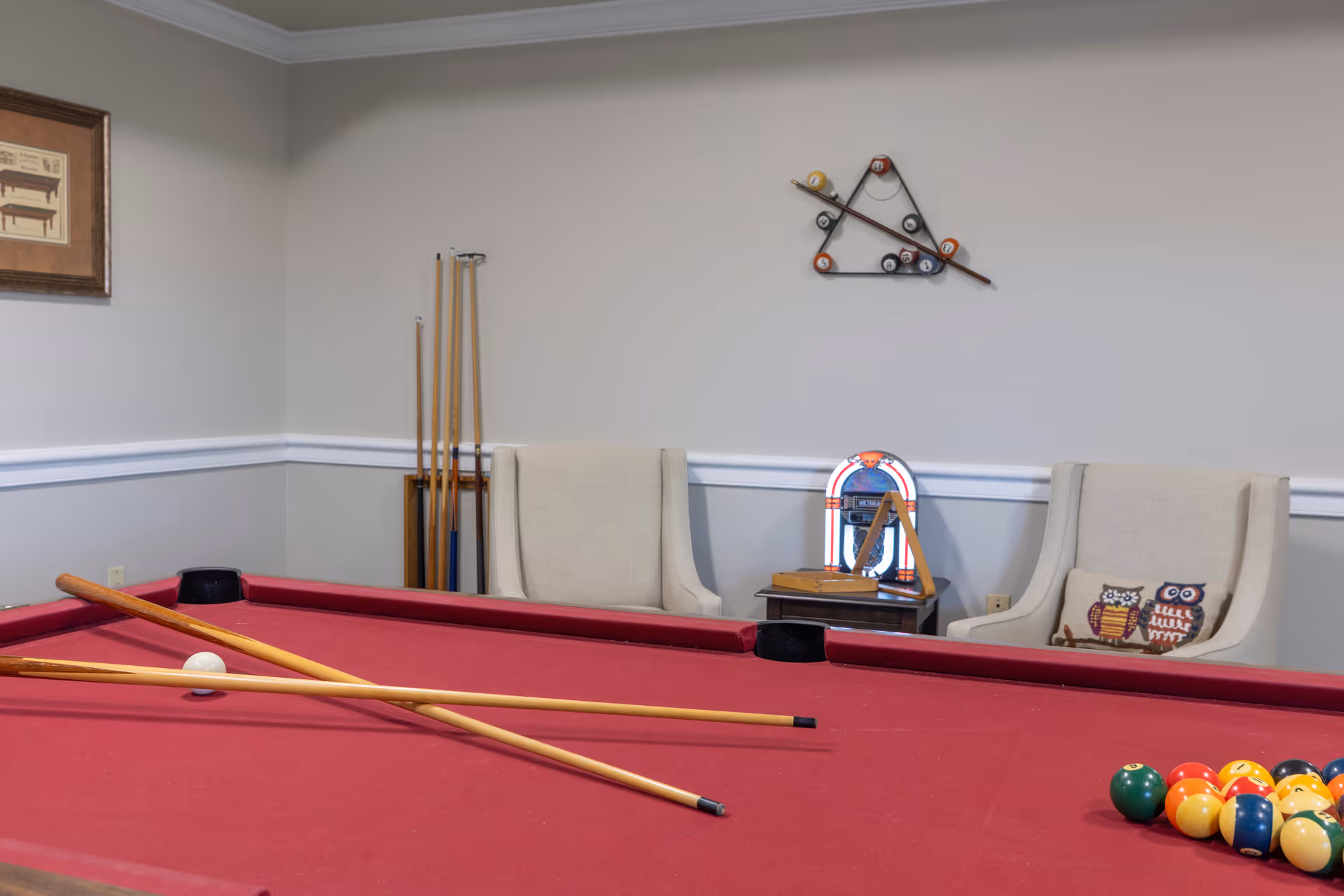 A recreational room with a red pool table in the foreground, two beige armchairs with owl-themed pillows, a small side table with a jukebox, and a wall-mounted rack holding pool cues and billiard balls arranged in a triangle.