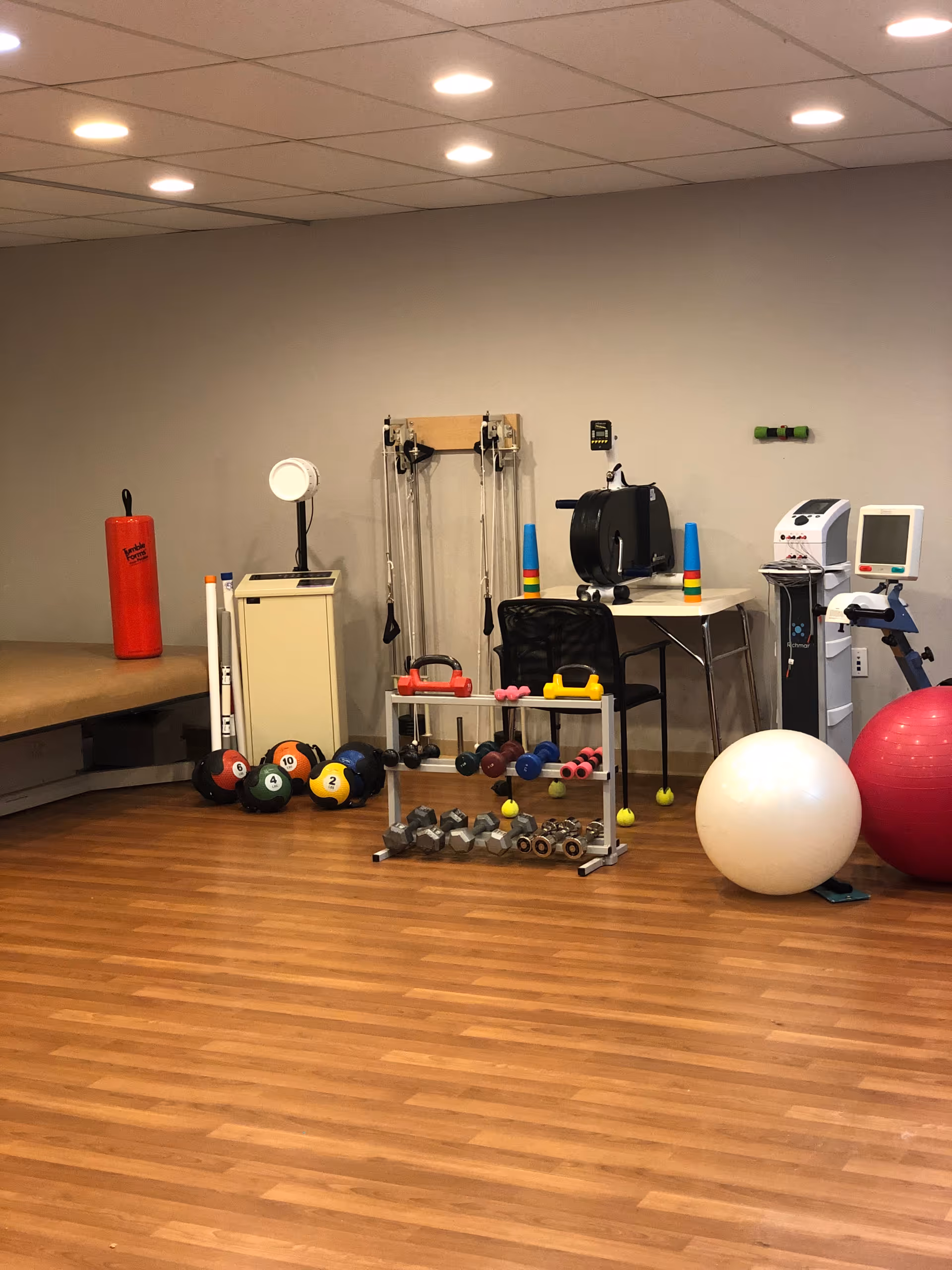 An indoor rehabilitation exercise room with dumbbells, medicine and stability balls, resistance equipment and other therapy gear on wood flooring.