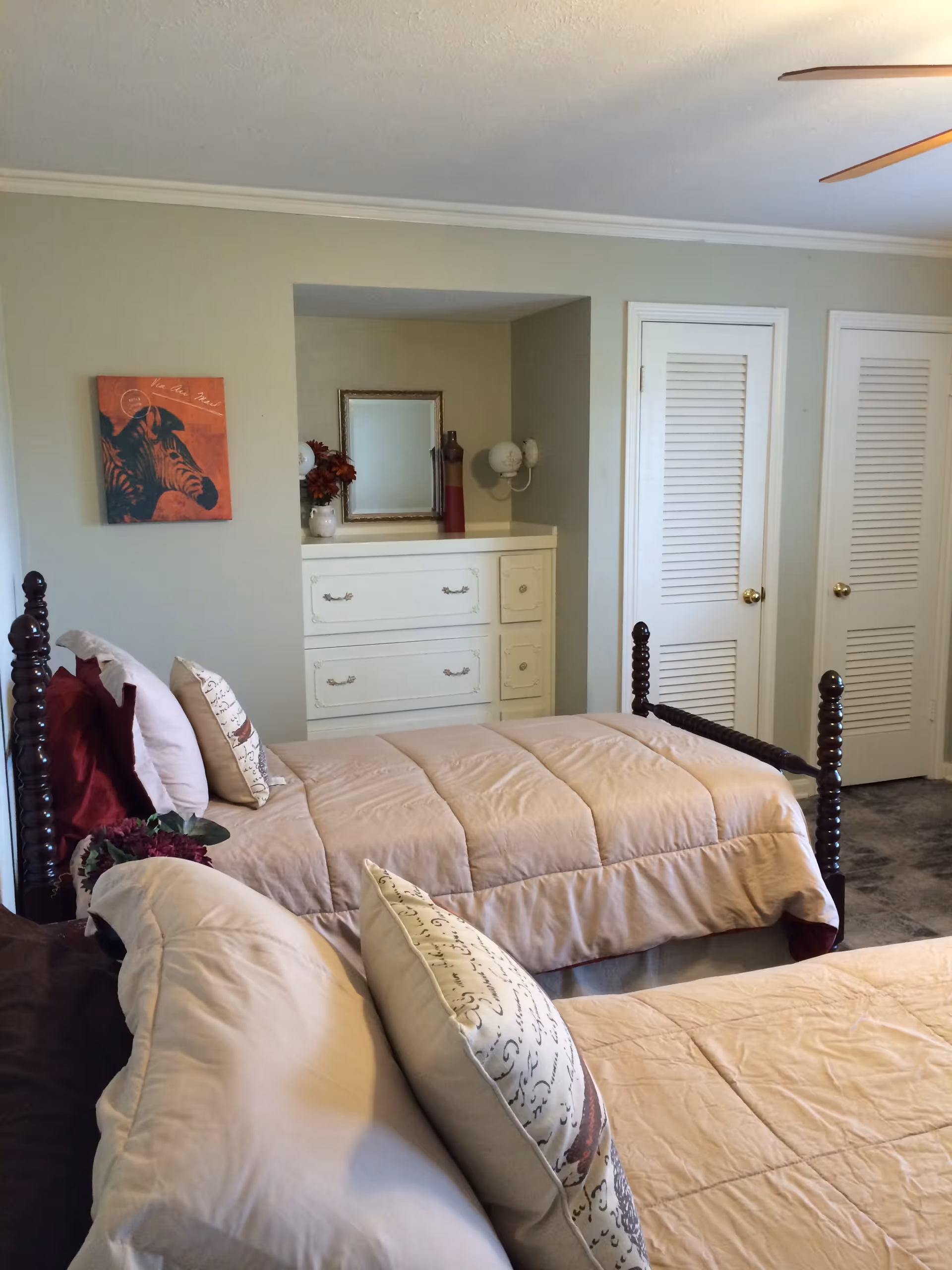 Two twin beds with beige comforters and decorative pillows in a bedroom with a dresser, mirror, and louvered closet doors.