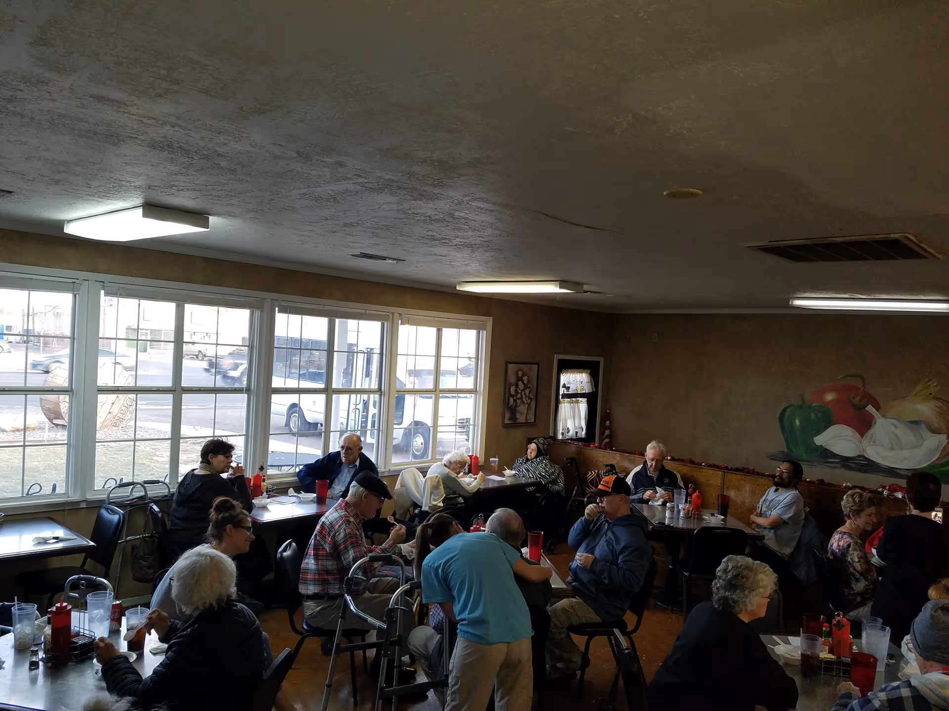 A group of elderly people sitting and socializing at tables in a dining area with large windows letting in natural light. The room has beige walls, a mural of vegetables on one wall, and red cups on the tables.