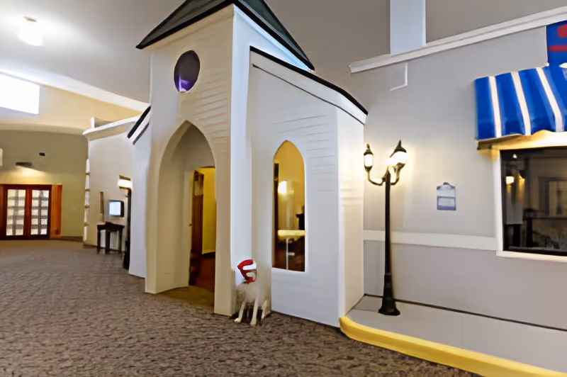 Interior view of a senior living facility hallway featuring a small white structure resembling a chapel or house with arched doorways and windows. A decorative dog statue wearing a red and white hat is placed near the entrance. The hallway has carpeted flooring, a tall lamp post, and a window with a blue and white striped awning.