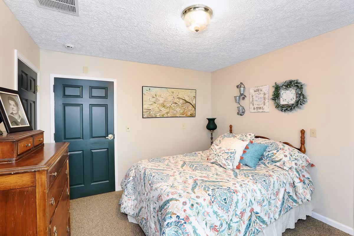 A cozy bedroom with a bed covered in a floral patterned comforter and multiple pillows. The walls are beige and decorated with a framed painting, a wreath, and other wall hangings. There is a wooden dresser with a framed black and white photo on top. The door is painted dark green.
